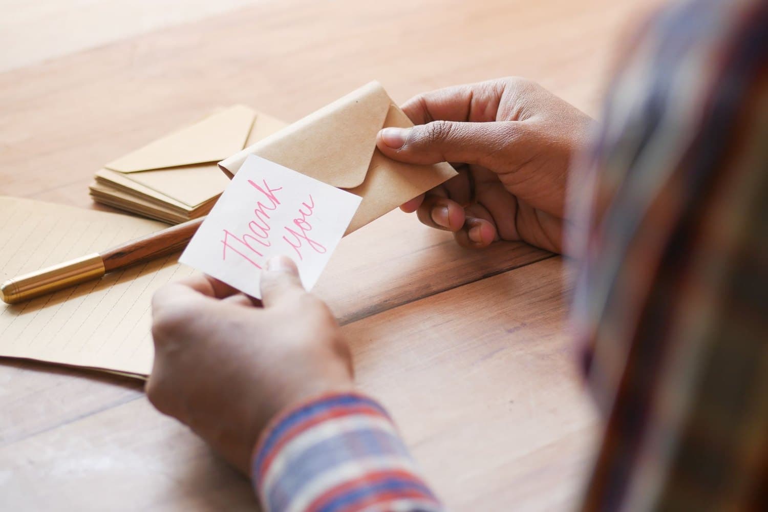 Hands holding a handwritten thank-you card and envelope at a wooden desk